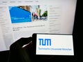 Person holding smartphone with logo of education institution Technical University of Munich on screen in front of web page.