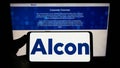 Person holding smartphone with logo of eye care company Alcon AG on screen in front of website.