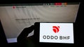 Person holding smartphone with logo of Franco-German financial company Oddo BHF AG on screen in front of website.