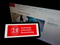 Person holding smartphone with logo of German university Technische UniversitÃÂ¤t Braunschweig on screen in front of webpage.