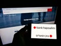 Person holding smartphone with logo of Israeli financial company Bank Hapoalim BM on screen in front of business webpage.