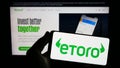 Person holding smartphone with logo of Israeli social trading company eToro on screen in front of website.