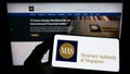 Person holding smartphone with logo of Monetary Authority of Singapore (MAS) on screen in front of website.