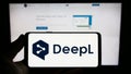Person holding smartphone with logo of neural machine translator DeepL on screen in front of website.