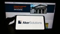 Person holding smartphone with logo of Norwegian engineering company Aker Solutions ASA on screen in front of website.