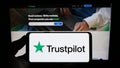 Person holding smartphone with logo of review platform company Trustpilot Group plc on screen in front of website.
