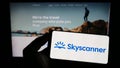 Person holding smartphone with logo of travel metasearch company Skyscanner Ltd. on screen in front of website.