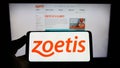 Person holding smartphone with logo of US animal drug company Zoetis Inc. on screen in front of website.