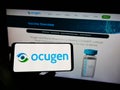 Person holding smartphone with logo of US biopharmaceutical company Ocugen Inc. on screen in front of website.