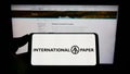 Person holding smartphone with logo of US company International Paper Company (IP) on screen in front of website.