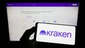 Person holding smartphone with logo of US crypto company Payward Inc. (Kraken) on screen in front of website.