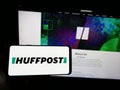 Person holding smartphone with logo of US news company HuffPost on screen in front of website.