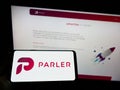 Person holding smartphone with logo of US social networking company Parler Inc. on screen in front of website.