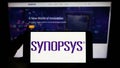 Person holding smartphone with logo of US technology company Synopsys Inc. on screen in front of website.