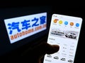 Person holding smartphone with web page of Chinese car trading platform Autohome Inc. on screen in front of logo.