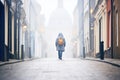 person in a hooded cloak walking away down a foggy, cobbled street