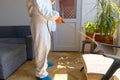 Person in protective suit with decontamination sprayer bottle disinfecting household and furniture