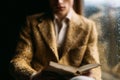 Person reading book while sitting by window with raindrops. Warm and inviting atmosphere highlighted by soft light and Royalty Free Stock Photo