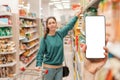 A person`s hand holds a mobile phone, with a shopping basket on the screen. In the background, a woman selects products in a