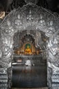 A person seated in front of an illuminated Buddha statue seen through a doorway decorated with silver sculptures.