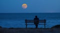 A person sits on a wooden bench facing the ocean the moon casting a soft glow over their face as they ponder their