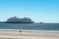 Person sitting on the shore of the beach during a beautiful sunny day, observing a large cruise ship anchored at a pier.
