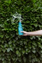 A person sprinkling water in a blue bottle with a plastic-free print in front of green bay laurels