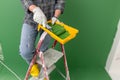 Person on a stepledder in working gloves holding yellow paint trough with middle-sized paint roller with green paint in front of