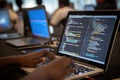 Person typing code on a laptop in a dimly lit room with other laptops and people in the background Royalty Free Stock Photo