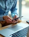 Person uses phone, laptop for two-factor authentication. Virtual security shield icon appears on phone screen. Modern tech