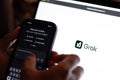 Tech guy trying out Grok in mobile phone with the laptop screen showing Grok logo, an generative, conversational, neural network,