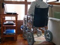 Person using a wheelchair, with a plaster cast on her leg, lloking through the window..Small desk with a laptop close to her.