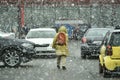 A person walks on the street among the cars coming and going from a parking lot during a snowstorm in early spring.