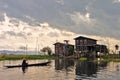 A person wearing a conical hat in a small boat rowing towards a stilted house during sunset at the Inle Lake in Myanmar. Royalty Free Stock Photo