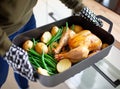 A person wearing oven mitts is holding an open roasting pan with green beans, potatoes, and chicken in the kitchen at