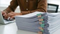 a person is working at a laptop, a stack of documents with colorful binder clips on a white table is nearby Royalty Free Stock Photo