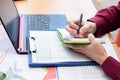 A person writes in a notebook data from a laptop and documents laid out on the table