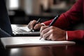 a person writing on a notebook and sitting near a laptop