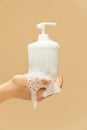 Personal Hygiene. Hand Covered With Foam Holding Liquid Soap Bottle On Beige Background. Washing Hands For COVID-19 Prevention