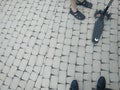 A persons feet shoes standing on grey cobblestone paving on a street and trotinet.