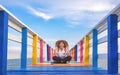 Happy Asian woman smiling while sitting on colorful wooden bridge at sea viewpoint against cloud on blue sky Royalty Free Stock Photo