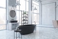 Perspective view on modern black bath in huge sunlit loft style bathroom with metallic furniture, round mirror on concrete wall Royalty Free Stock Photo