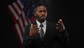 Persuasive confident African American man talking and gesturing with blurred US flag at black background. Portrait of