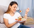 Peruvian housewoman eating vegetable salad on sofa at home