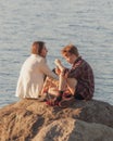 A pair of teenagers sits opposite each other on a large stone on the shore Royalty Free Stock Photo
