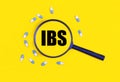 Pharmaceutical tablets and capsules near a magnifying glass with the text IBS Irritable Bowel Syndrome on a yellow background.