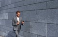 With phone and drink in hands. Young successful businessman in grey formal wear is outdoors in the city