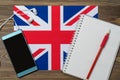 A phone, headphones, spiral notebook, and red pencil rest on the flag of the United Kingdom. Brown wooden background