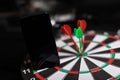 Phone stands on Darts field with Three Darts to hit red target close-up