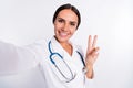 Photo of adorable cheerful young woman doctor wear formal coat tacking selfie smiling showing v-sign isolated white
